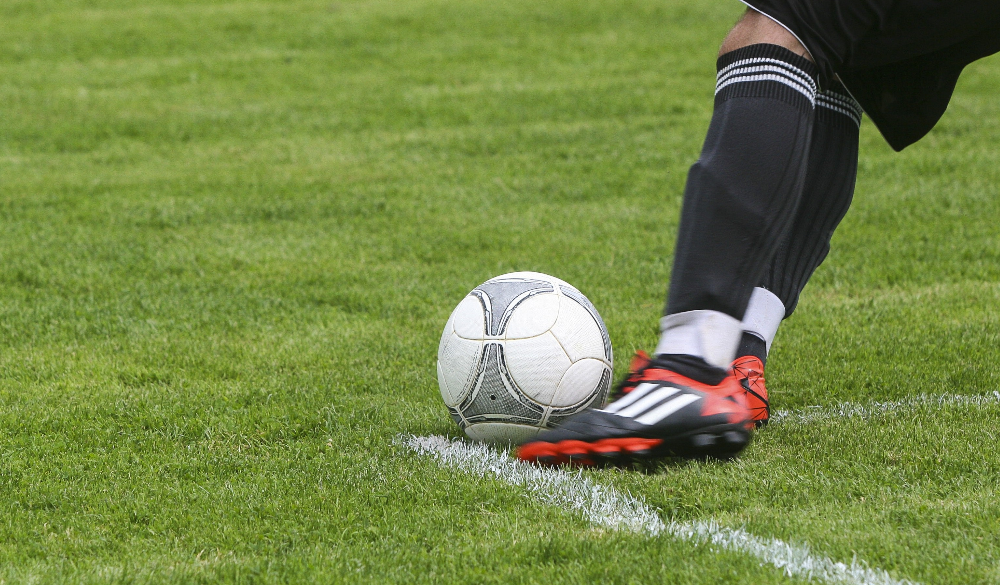 Young footballer in orange and black cleats kicks white football on a grass field. YDFL Presentation