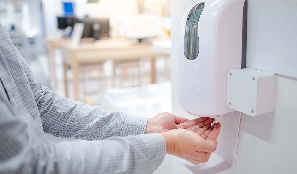 Male hands using automatic hand sanitiser dispensers for hand hygiene in an office.