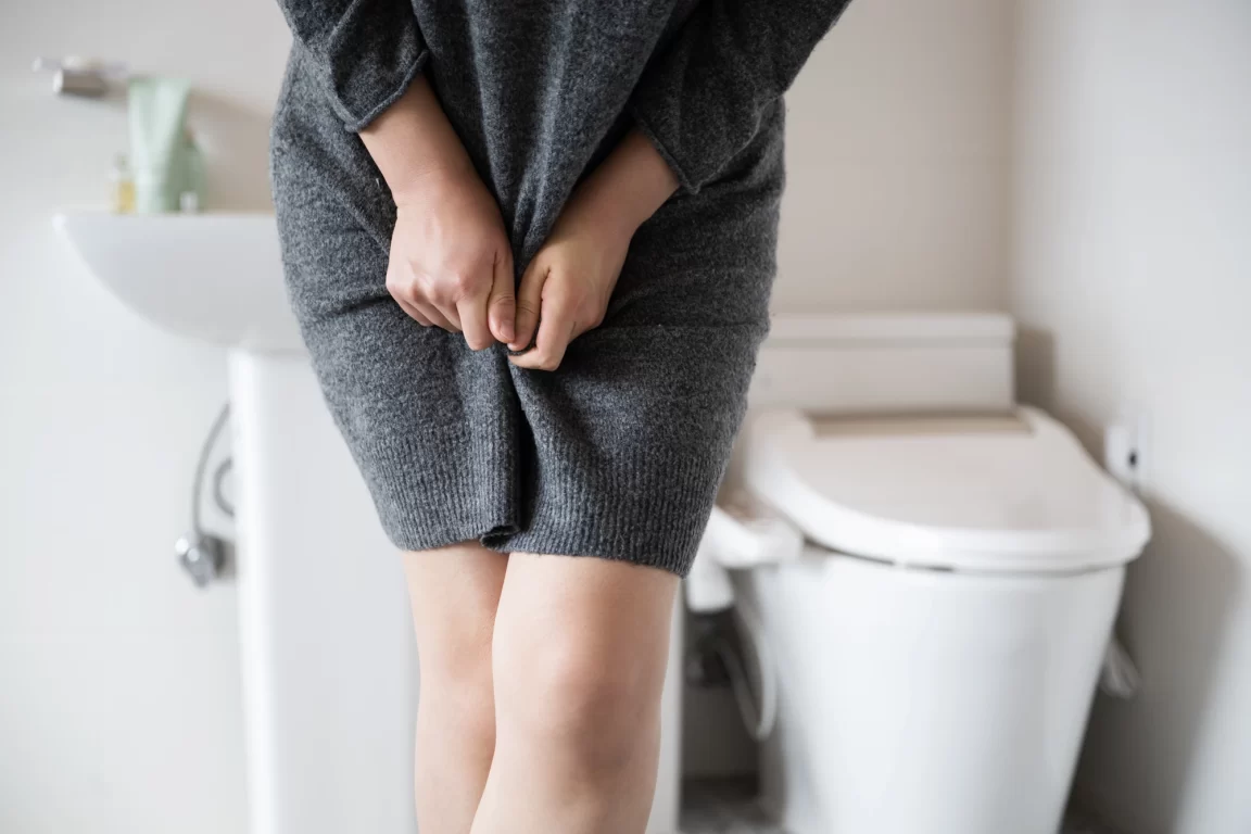 Woman in bathroom in grey knitwear crosses legs, suffering from incontinence.