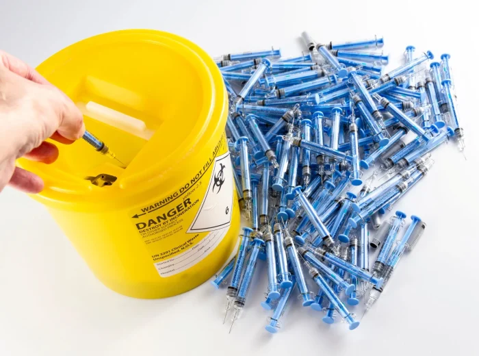 Yellow biohazard sharps disposal container, partially filled, with blue needles next to it.