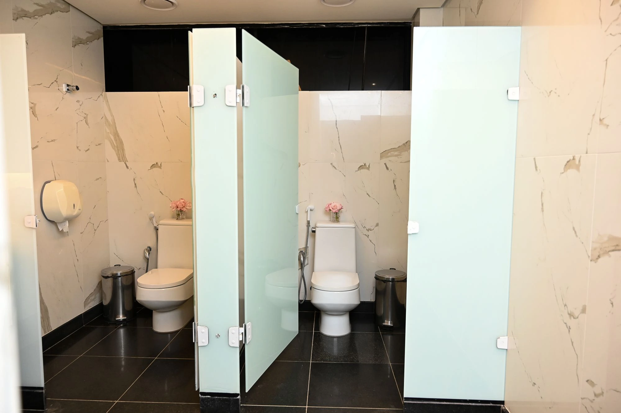 Two modern, marble-tiled bathroom stalls with frosted glass doors, each containing a white toilet, a stainless steel sanitary bin, and a small vase of pink flowers.