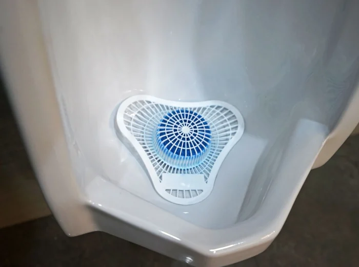 A white, triangular plastic urinal screen with a blue deodorising block in the centre, placed inside a clean white urinal.