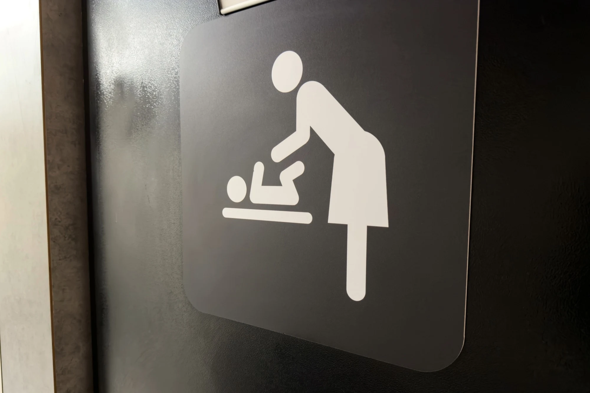 Grey washroom sign featuring a white pictogram of a person changing a baby's nappy.