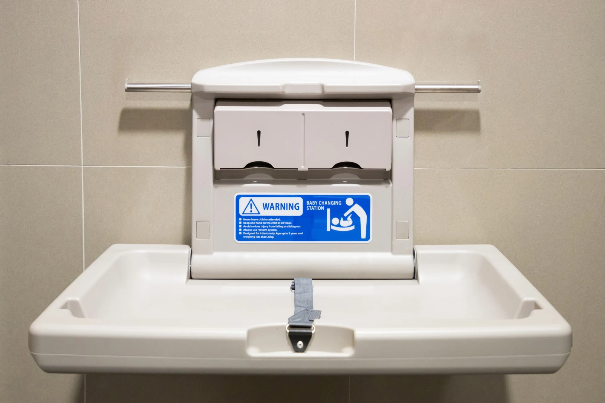 A wall-mounted, beige plastic baby changing station folded open, featuring a grey safety strap and a blue warning label.